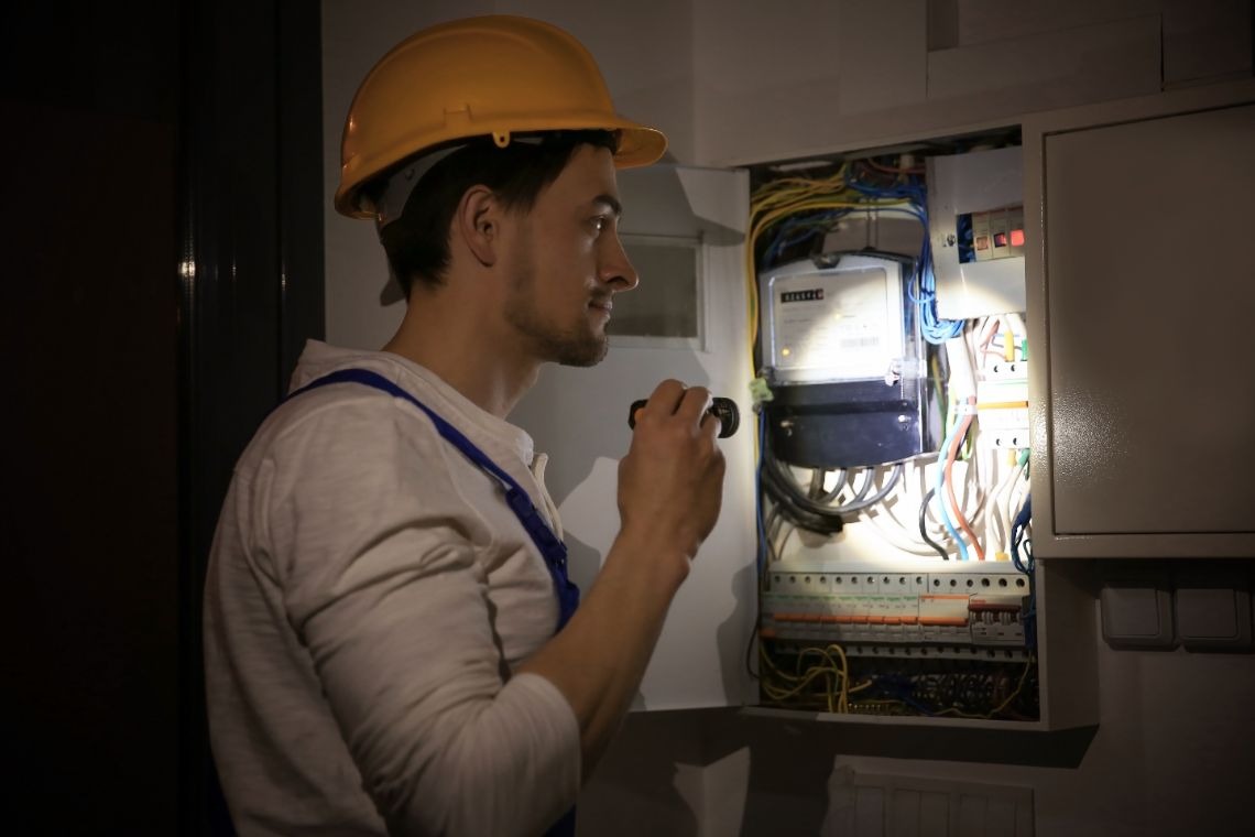 Elektryk używa lampy roboczej budowlanej w formie latarki, aby sprawdzić bezpieczniki